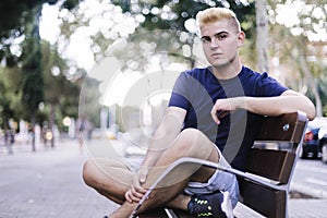 Portrait of a young teenage male with blonde bangs sitting on a bench in the street while looking at the camera