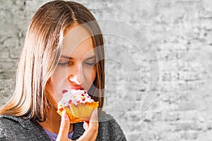 Young teenager brunette girl with long hair eating cream cake on gray wall background