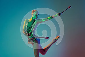 Portrait of young tender girl, female rhythmic gymnast performing with clubs isolated over gradient blue background in
