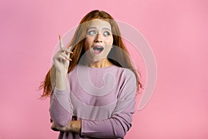 Portrait of young thinking pondering woman having idea moment pointing finger up on pink studio background. Smiling