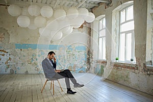 Portrait of young thoughtful man, businessman sitting on chair with laptop in empty room. Choosing building for company