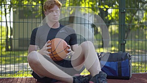 Portrait of young thoughtful redhead man sitting with ball on basketball court. Sad Caucasian lonely sportsman after