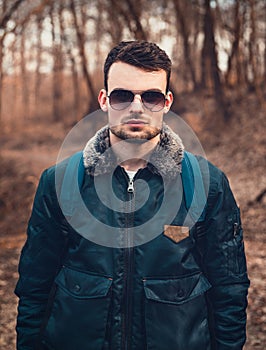 Portrait of young trendy man in blue aviator bomber jacket with sunglasses  standing on the path in forest.  Fashion male model