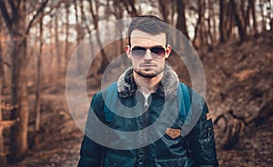 Portrait of young trendy man in blue aviator bomber jacket with sunglasses  standing on the path in forest.  Fashion male model