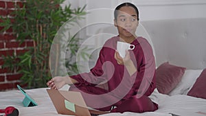 Portrait of young troubled woman sitting on bed holding coffee cup thinking. Restless beautiful African American