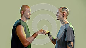 Portrait of young twin brothers with tattoos and piercings looking at each other, giving fist bump, standing face to