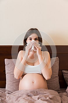 Portrait of young unhappy pregnant woman leaning on pillows on bed and blowing her nose into tissue. Future mom caught cold and