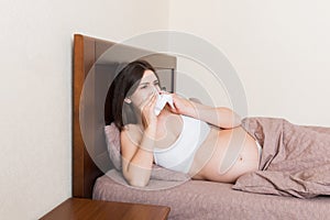 Portrait of young unhappy pregnant woman leaning on pillows on bed and blowing her nose into tissue. Future mom caught cold and