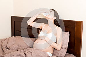 Portrait of young unhappy pregnant woman leaning on pillows on bed and blowing her nose into tissue. Future mom caught cold and