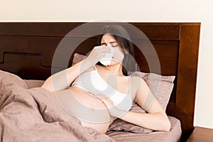 Portrait of young unhappy pregnant woman leaning on pillows on bed and blowing her nose into tissue. Future mom caught cold and