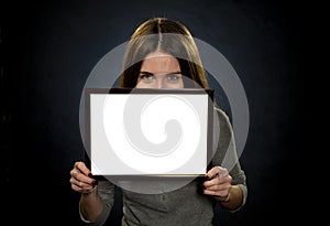 Portrait of a young woman 25-30 years old holding a white text frame on a dark background, shyly hiding her face, smiling