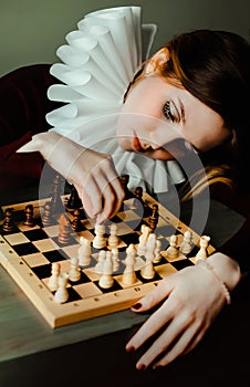 Portrait of an young woman in an antique dress with a white frill, who is thinking about the next move in a chess game. The queen