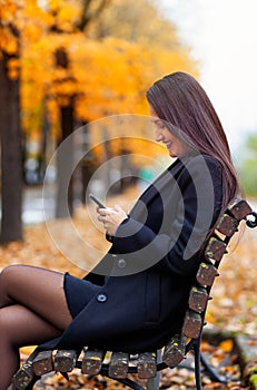 Portrait of a young woman in autumn. She smiles using the smartphone