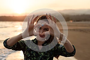 Portrait of a young woman on the beach at red sunset, heart of fingers, message of love