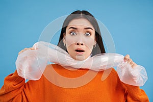 Portrait of young woman being choked by plastic bags, concept of recycling and environment, blue background