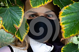 Portrait of the young woman in a black protective mask with a framed face of natural tree leaves.