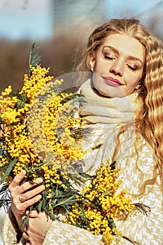 Portrait of a young woman with blond hair holding a bouquet of mimosa in her hands. Spring