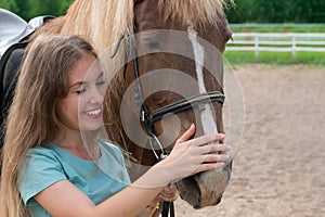 Portrait of young woman with blond long hair and smile is caressing brown horse snoot with big nostrils.