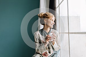 Portrait of young woman with blonde hair drinking coffee or tea next to big window, smiling, enjoying happy morning at
