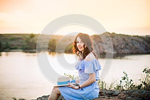 Portrait of young woman in blue romantic dress with hat sitting near river and enjoying sunset evening. Calm and harmony. Summer