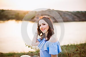 Portrait of young woman in blue romantic dress with hat sitting near river and enjoying sunset evening. Calm and harmony. Summer