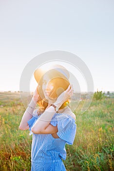 Portrait of young woman in blue romantic dress and straw hat on green meadow enjoying summer sunset. Calm and harmony. Summer