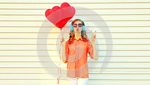 Portrait of young woman with bunch of red heart shaped balloons listening to music in headphones and blowing her lips sending