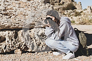 Portrait of young woman with camera. Sitting tourist taking photo of ancient ruins on sunny day. Blurred background