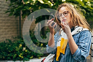 Portrait of a young woman in a casual outfit puting liptisck on her lips, posing outdoors. Pretty female student with long red