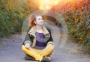 Portrait with a young woman closed her eyes and relaxed in the lotus position sitting on an asphalt path in an autumn park. Calm