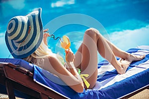 Young woman with cocktail glass chilling in the tropical sun near swimming pool on a deck chair