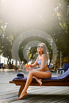 Portrait of young woman with cocktail glass chilling in the tropical sun near swimming pool on a deck chair with palm