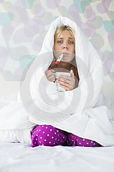 Portrait of young woman with coffee mug taking temperature while wrapped in quilt on bed