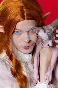 Portrait of young woman cosplay elf with red hair in white dress holding half asleep Sphinx kitten