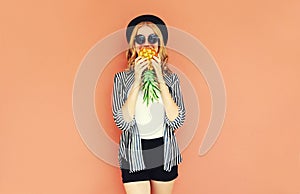 Portrait of young woman covering her mouth with pineapple wearing a black hat, sunglasses, striped shirt on background