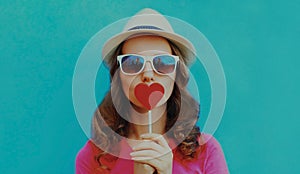 Portrait young woman covering her mouth with red heart shaped lollipop on a blue background