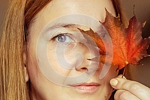 Portrait of a young woman covering one eye with an autumn fallen maple leaf. Autumn mood