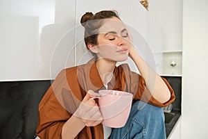 Portrait of young woman with cup of coffee, sits in kitchen and drinks aromatic drink at home, holds tea mug