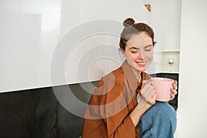 Portrait of young woman with cup of coffee, sits in kitchen and drinks aromatic drink at home, holds tea mug