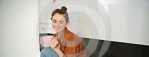 Portrait of young woman with cup of coffee, sits in kitchen and drinks aromatic drink at home, holds tea mug