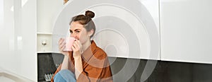 Portrait of young woman with cup of coffee, sits in kitchen and drinks aromatic drink at home, holds tea mug