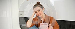 Portrait of young woman with cup of coffee, sits in kitchen and drinks aromatic drink at home, holds tea mug