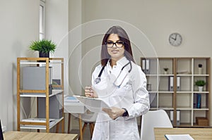 Portrait of young woman doctor in white coat standing in staff room and holding clipboard