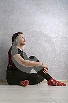 Portrait of young woman with dreadlocks sitting on the floor and thinking against gray background. Vertical frame