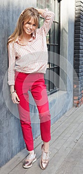 Portrait of a young woman dressed in a blouse, red Chino Trouser, and gold brogues. Posing next to a gray wall. Fashion. photo