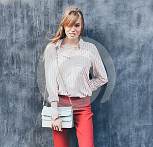 Portrait of a young woman dressed in a blouse, red Chino Trouser, a handbag turquoise on her shoulder. Posing next to a gray wall.
