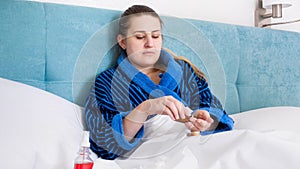 Portrait of young woman feeling unwell lying in bed and taking pills from bottle