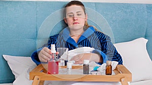 Portrait of young woman feeling unwell lying in bed and taking sore throat spray from wooden tray