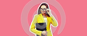 Portrait of young woman with folder wearing eyeglasses, yellow business suit on pink background, magazine style