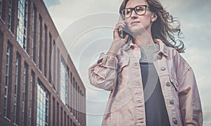 Portrait of young woman in glasses standing outdoors and talking on cellphone.Girl is walking along city street